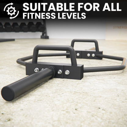 Black hex trap bar with curved handles and metal attachment points, displayed on a light concrete gym floor with dumbbells in the background.