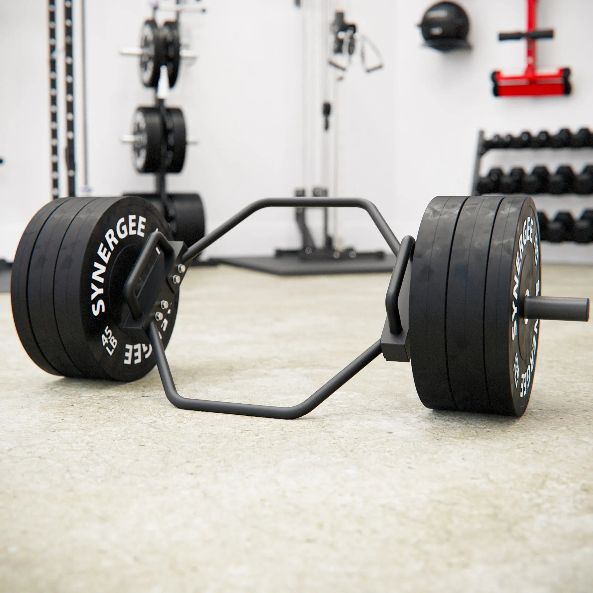 Hex trap bar with black rubber-coated weight plates loaded on each end, resting on a gym floor with various equipment visible in the background.