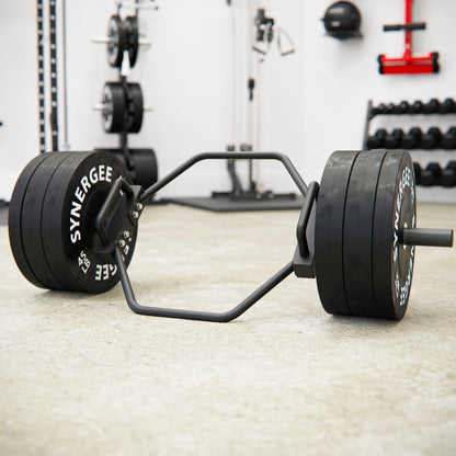 Hex trap bar with black rubber-coated weight plates loaded on each end, resting on a gym floor with various equipment visible in the background.