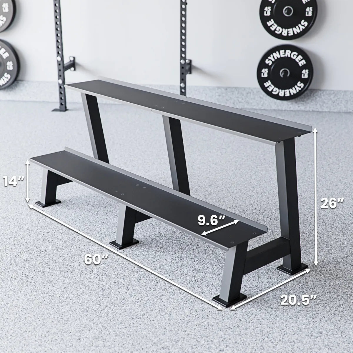 Two-tier black weight bench with metal legs positioned in a gym setting with a power rack and loaded plates visible in background. Dimensions labeled: 60" length, 26" height, 20.5" depth, and 9.6" seat width.