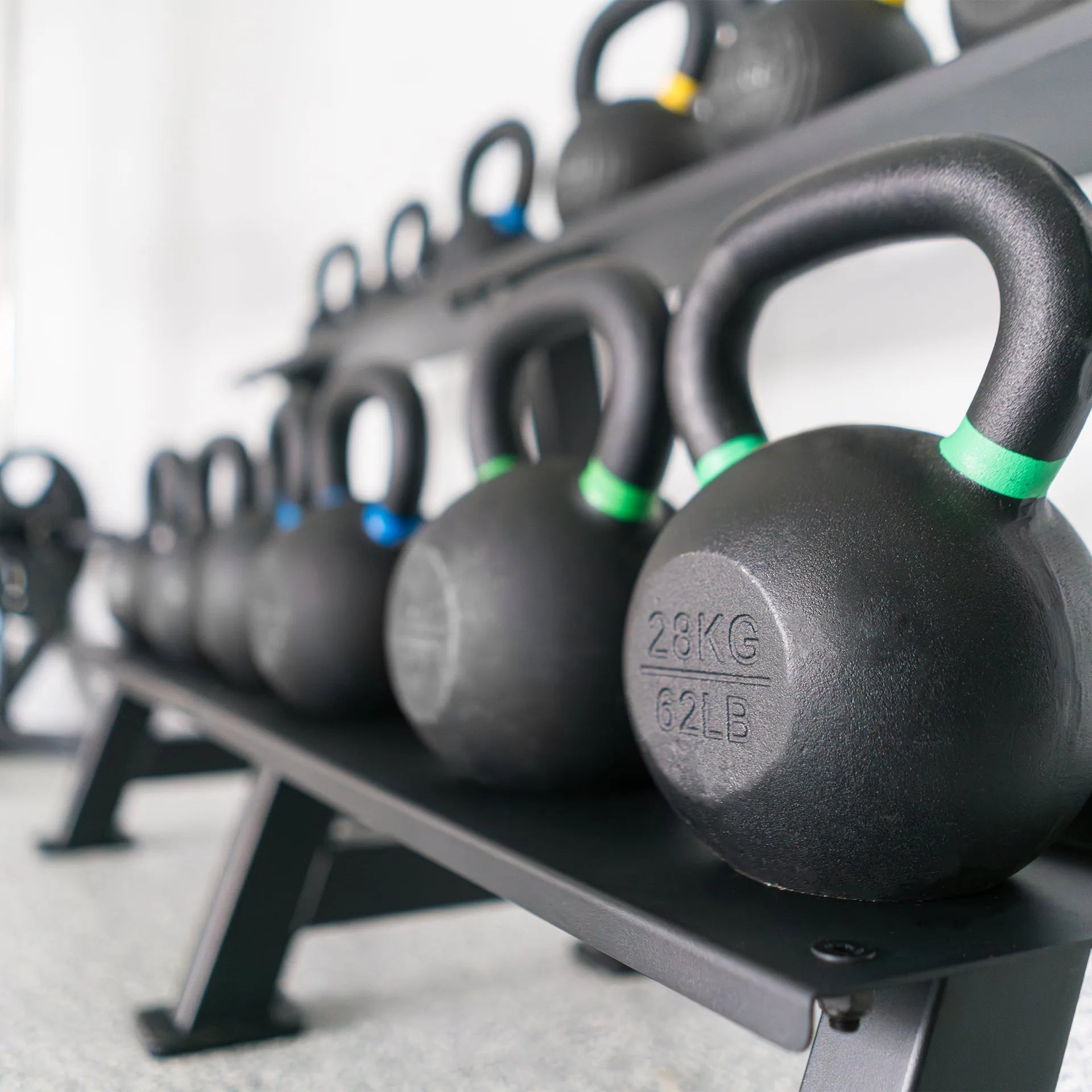 Row of black kettlebells with colored bands displayed on a gray storage rack, with the front kettlebell marked 28KG and 62LB.