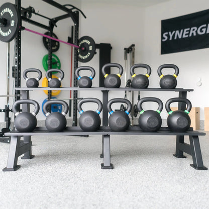 Two-tier black kettlebell storage rack holding twelve kettlebells with colored handles in varying weights, positioned in a gym with exercise equipment visible in the background.