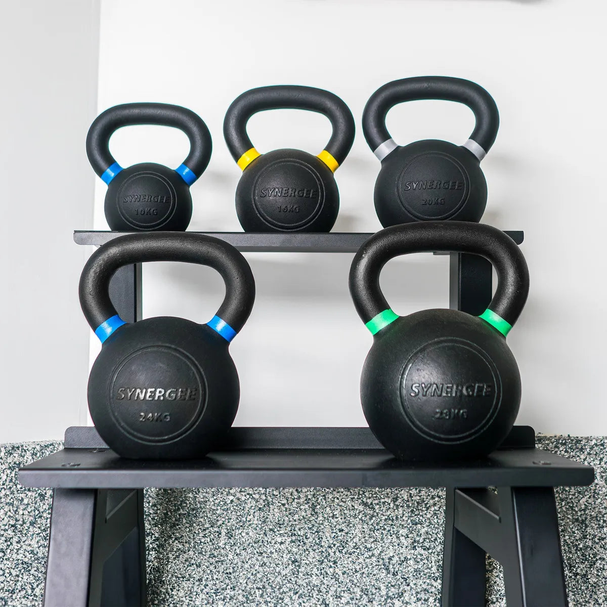 Five black cast-iron kettlebells with colored accent bands stored on a two-tier gray metal rack. Weight markings visible on each bell ranging from 10kg to 28kg.