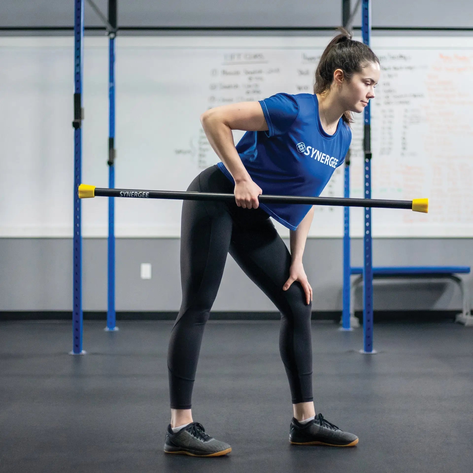 Woman performing a bent-over row exercise with a black weighted bar featuring yellow end caps, wearing a blue Synergee shirt in a gym setting.