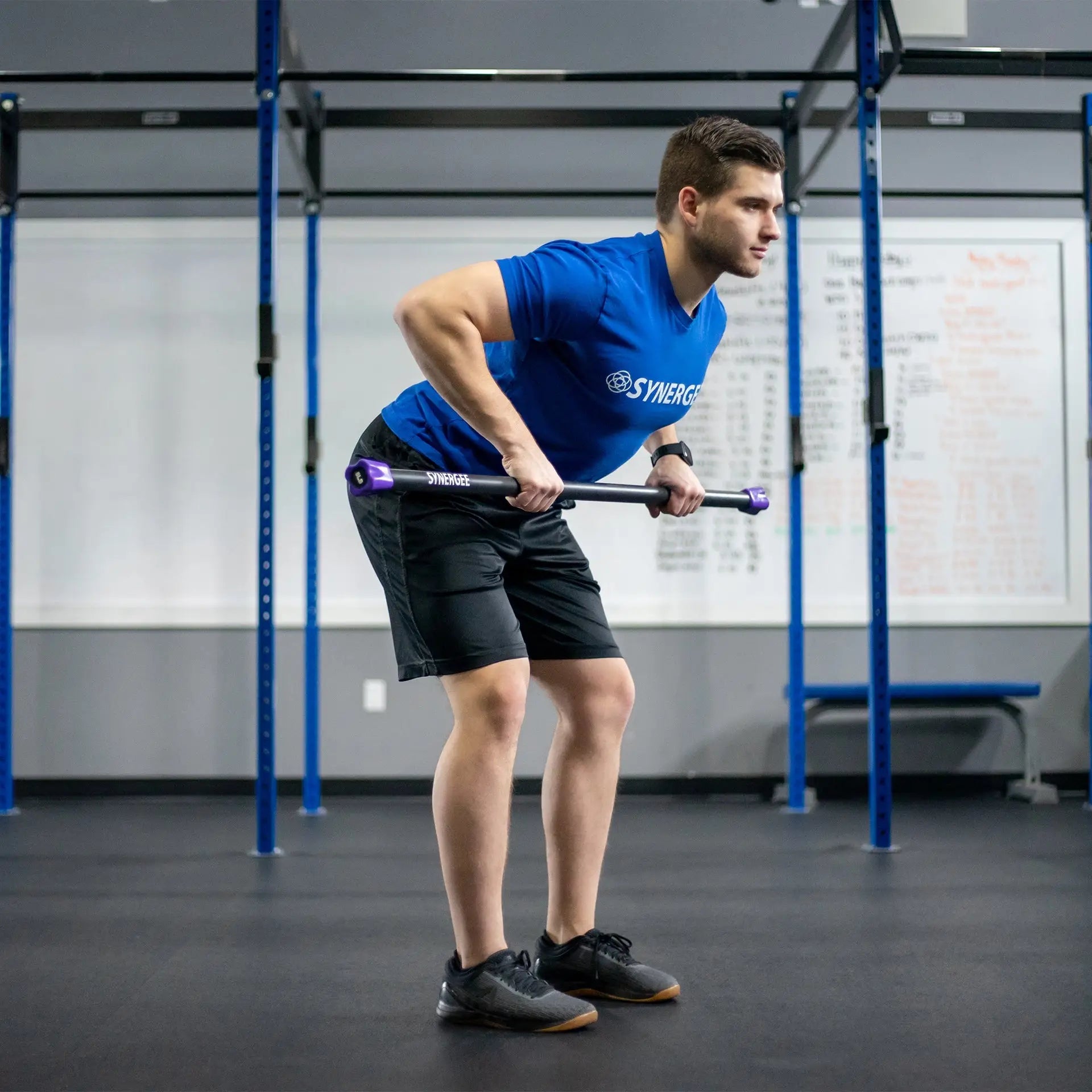 Man performing a bent-over row with a black weighted bar featuring purple end caps inside a CrossFit gym with blue rig frame.