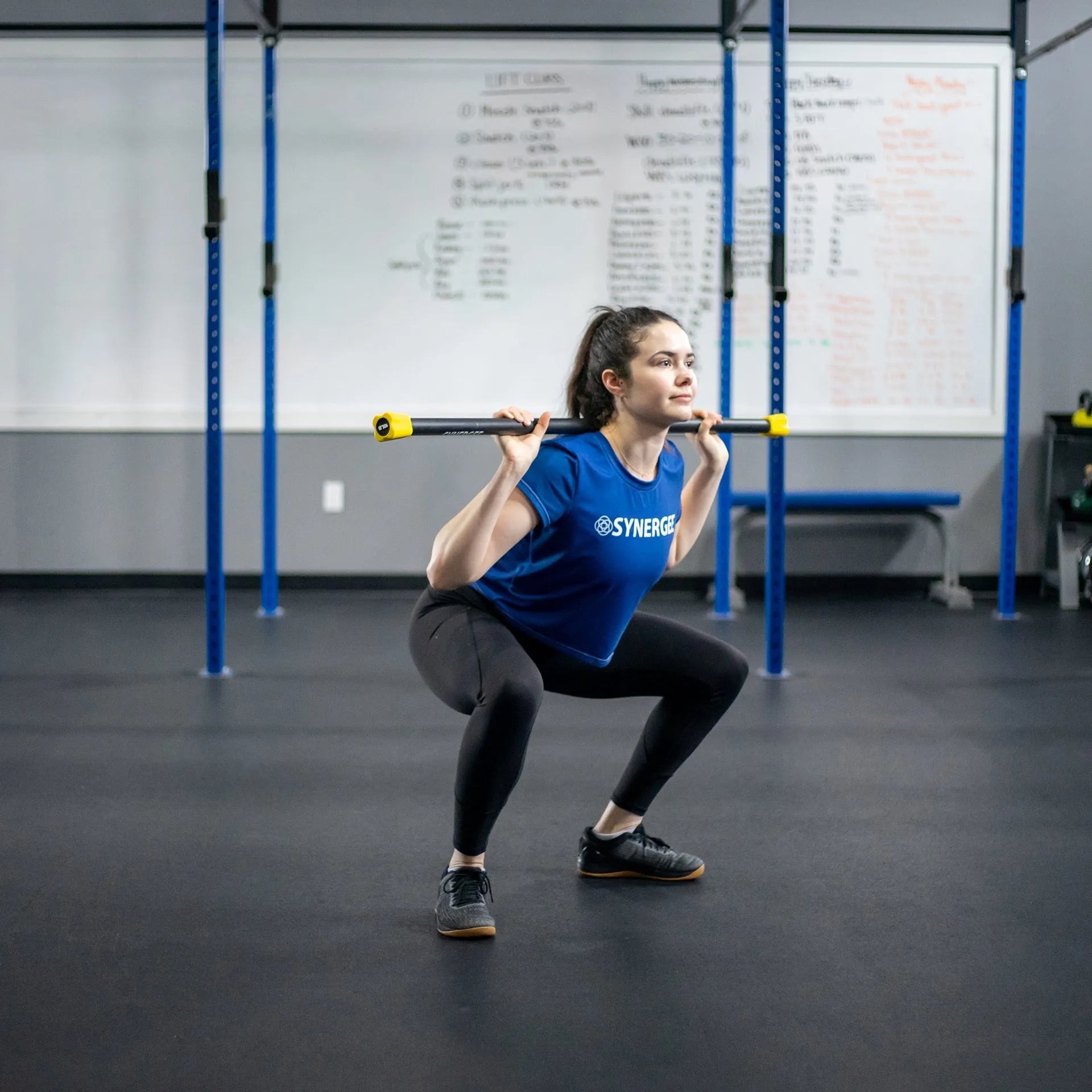Woman performing a back squat with a black and yellow weighted barbell across her shoulders in a CrossFit gym with blue rig uprights.