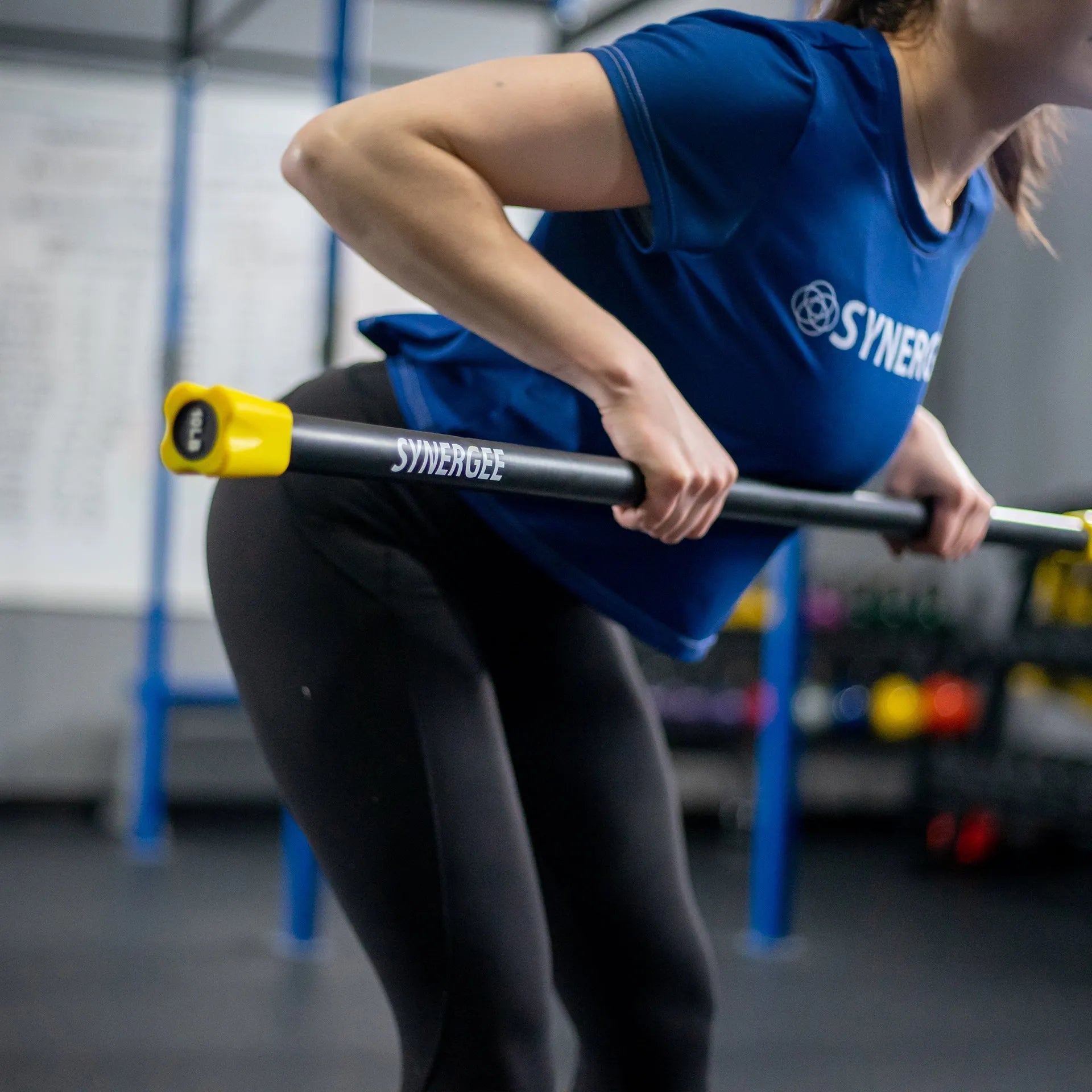 Woman performing a barbell exercise with a black Synergee weighted bar with yellow end caps in a gym setting.