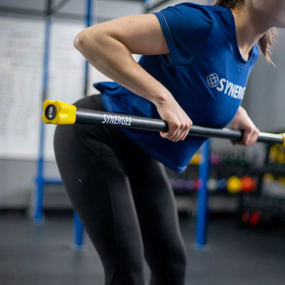 Woman performing a barbell exercise with a black Synergee weighted bar with yellow end caps in a gym setting.