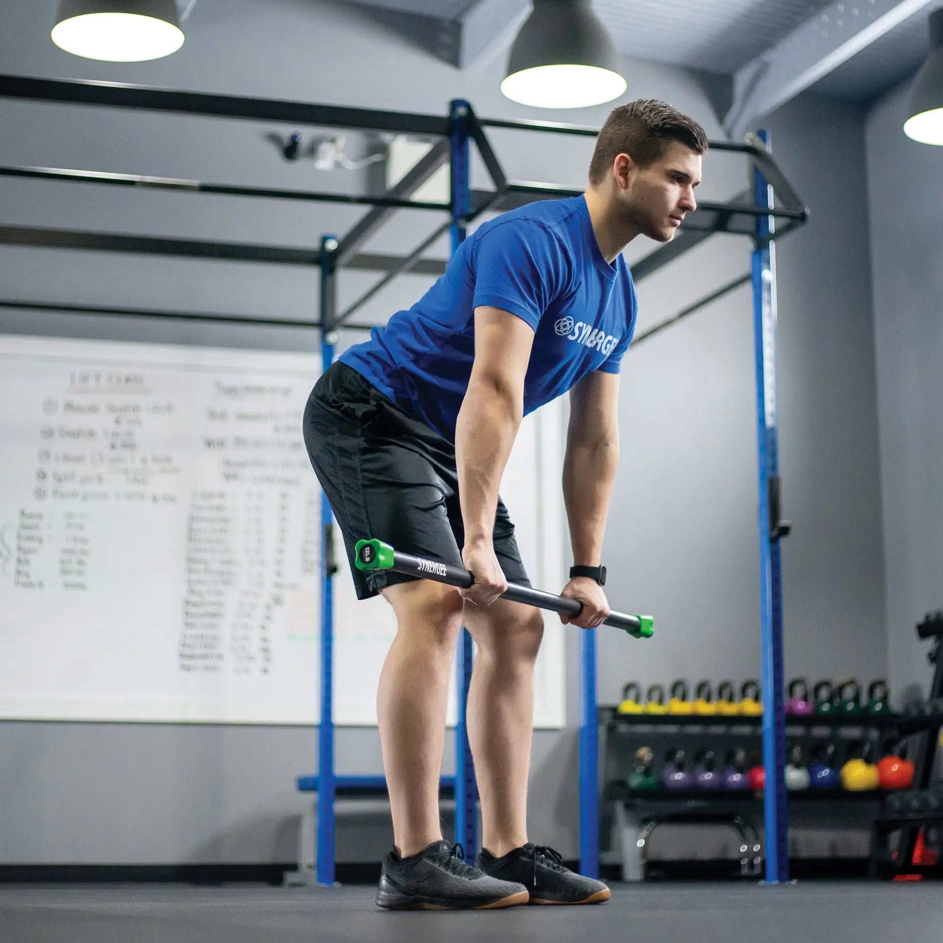 Man performing bent-over row exercise with a weighted bar featuring green end caps in a CrossFit-style gym with blue rig frame and dumbbell rack visible.