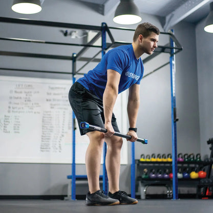 Man performing a bent-over rowing exercise with a weighted bar in a gym setting with blue rig frame and colorful medicine balls visible in the background.