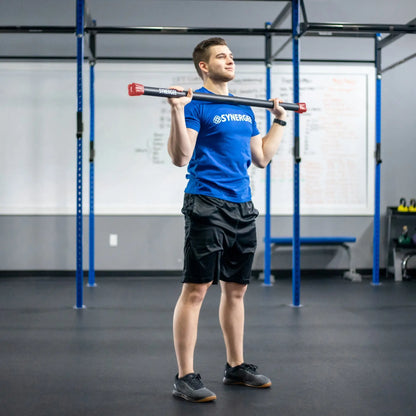 Man performing a shoulder press with a black weighted bar with red end caps inside a blue steel rig in a crossfit gym setting.