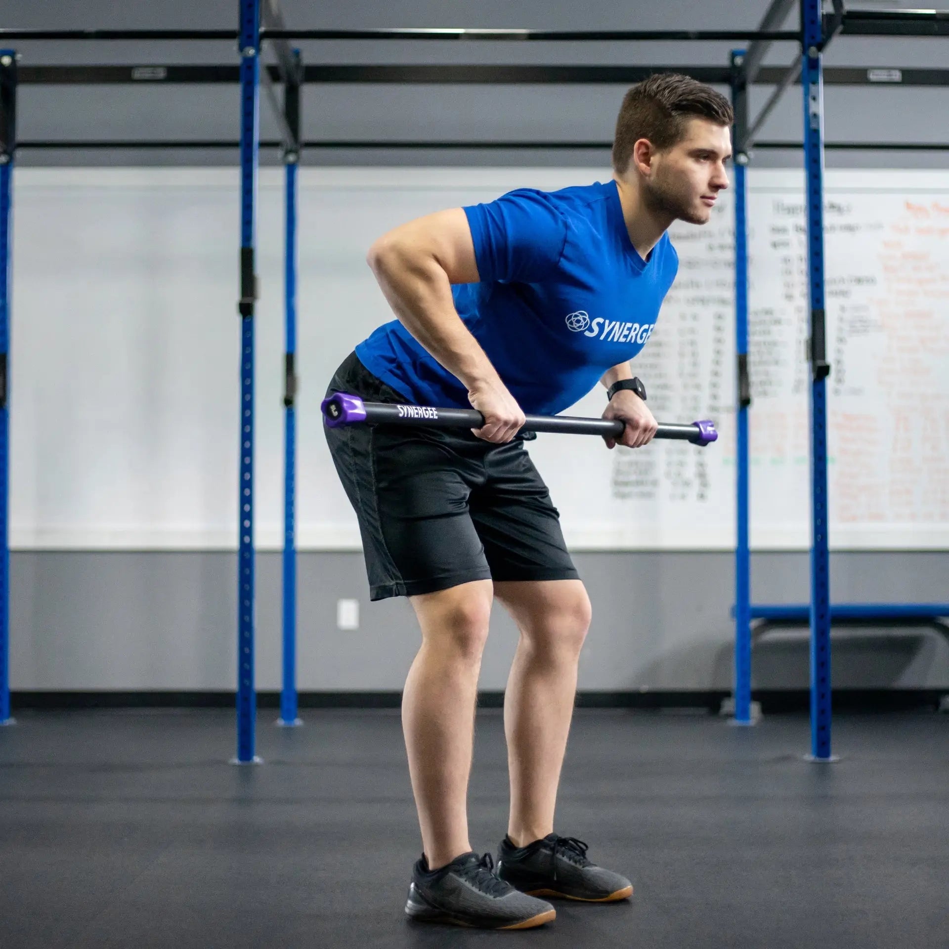 Man performing a bent-over row with a black weighted bar with purple end caps inside a blue crossfit rig in a gym setting.
