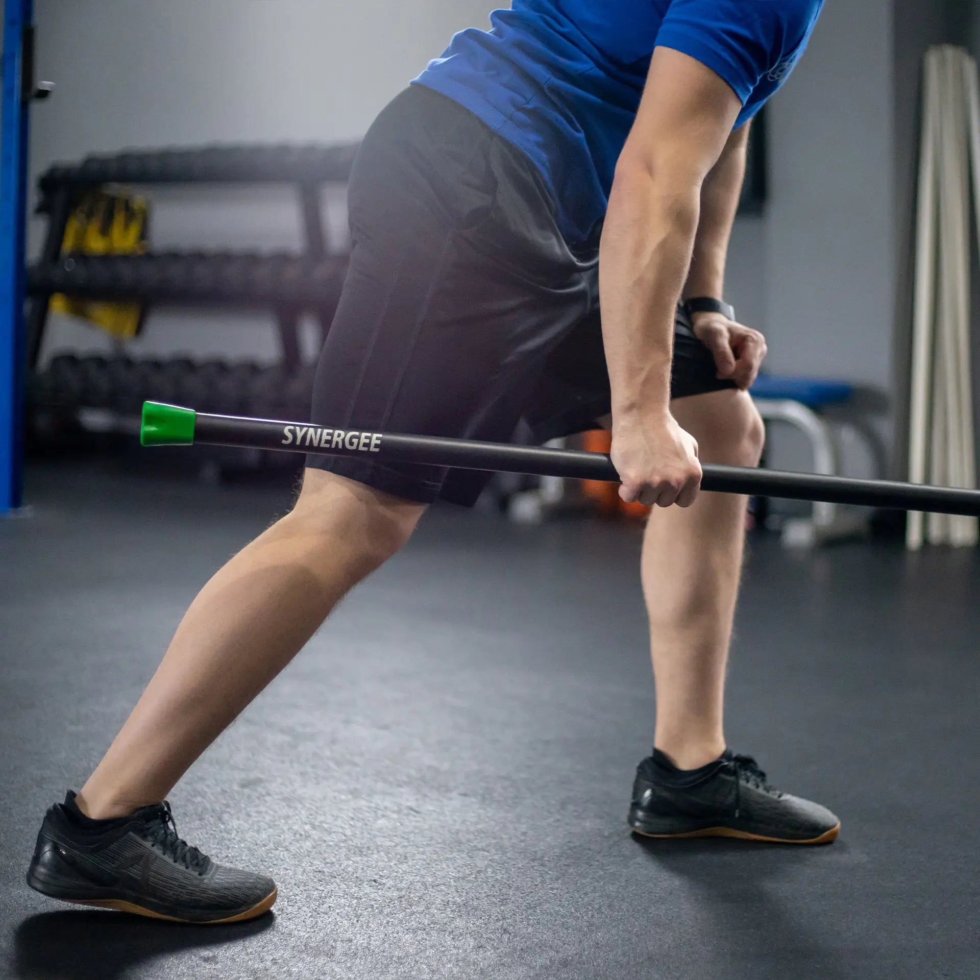 Man holding a black Synergee weighted workout bar with green and orange end caps during an exercise in a gym setting.