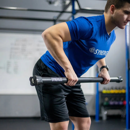 Man performing a bent-over row exercise with a black weighted bar labeled Synergee in a gym with blue rig structure and white walls.