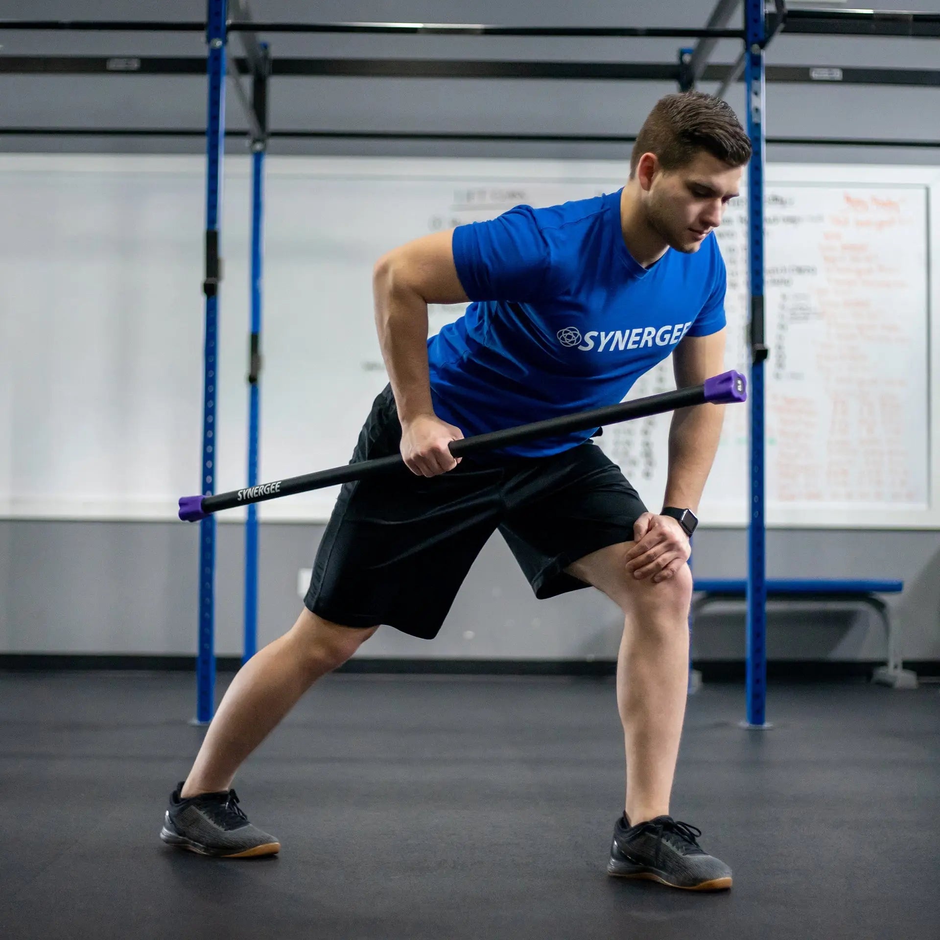 Man performing a lunge exercise while holding a black weighted bar with purple handles in a gym setting with blue rig frame.