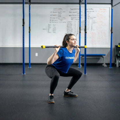 Woman performing a front squat with a weighted bar featuring yellow end caps in a CrossFit gym with blue rig uprights and whiteboard in background.