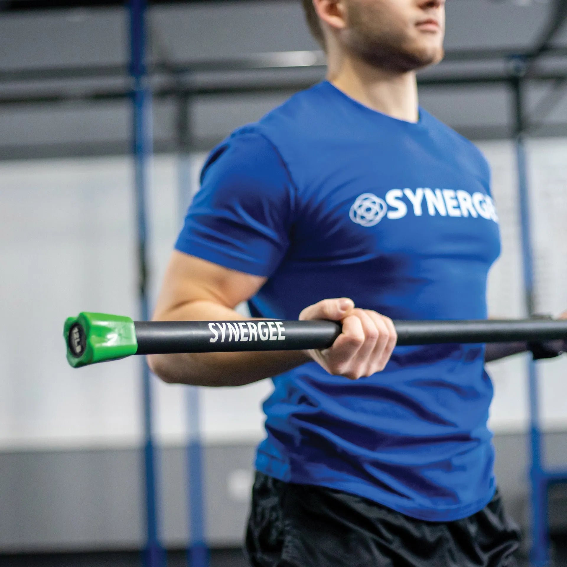 Man holding a black weighted bar with green end caps labeled Synergee in a gym setting with blue rig structure in background.