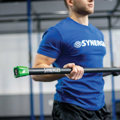 Man holding a black weighted bar with green end caps labeled Synergee in a gym setting with blue rig structure in background.