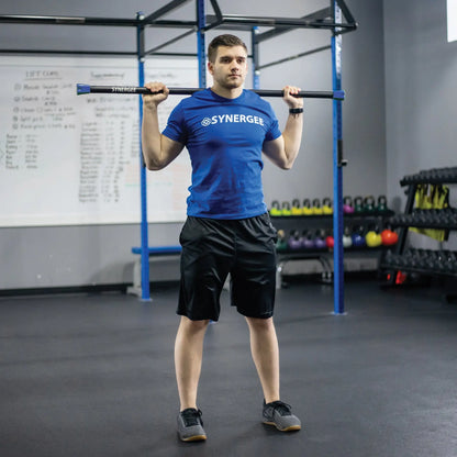 Man performing an overhead hold with a black weighted bar on a blue pull-up rig in a gym with dumbbells and kettlebells visible in the background.