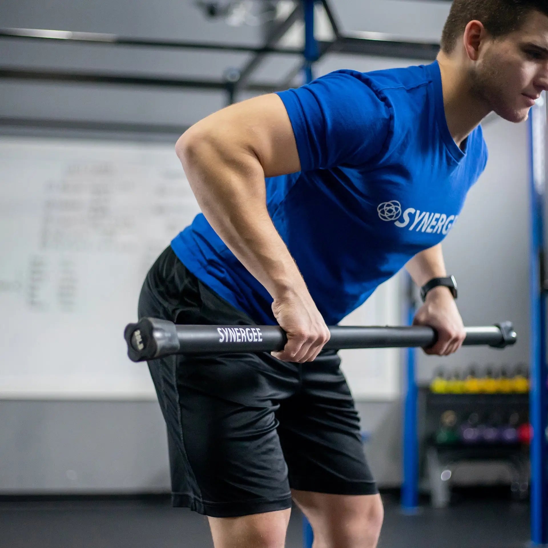 Man performing a bent-over row exercise with a black weighted bar labeled Synergee in a gym setting with blue rig structure in background.