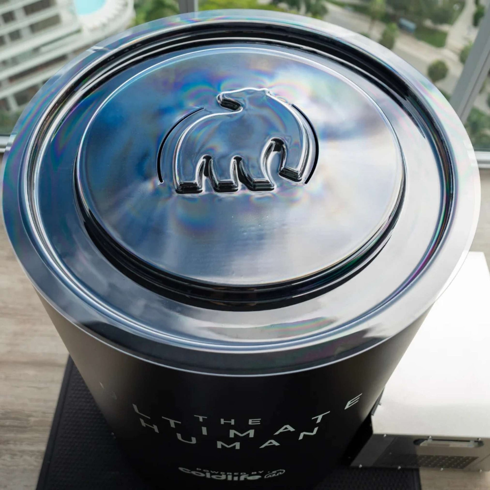 A close-up view of a sleek, metallic container with a bear logo on the lid, labeled THE ULTIMATE HUMAN and featuring the branding powered by Coolidge. The container is set against a backdrop of greenery and buildings visible through a window.