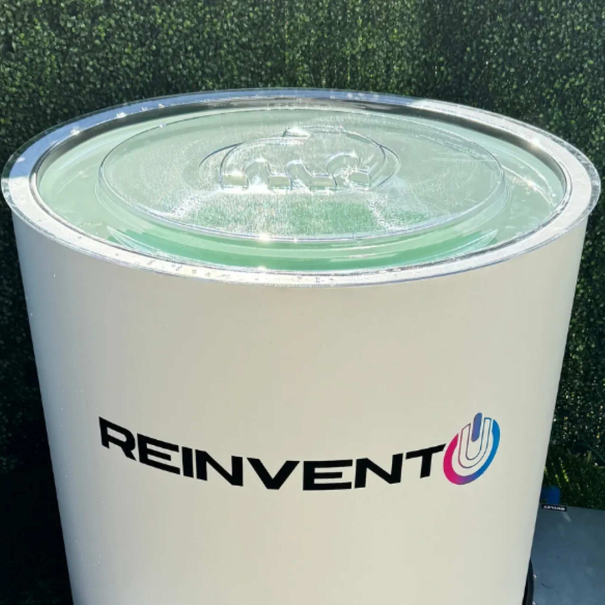 A cylindrical container with a clear top, featuring the logo REINVENT prominently displayed on its side, set against a green backdrop.