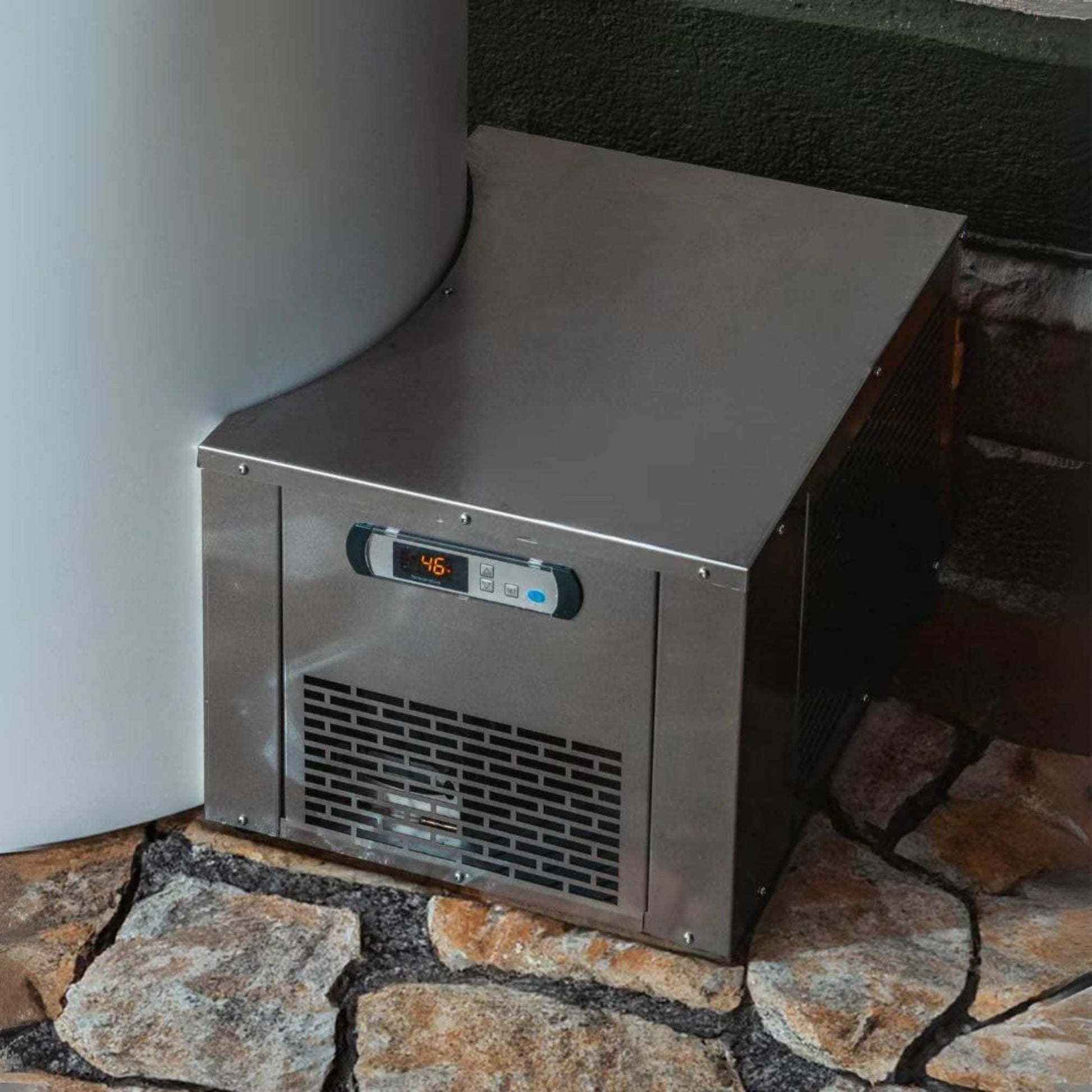 A stainless steel ice maker unit with a digital display showing a temperature of 46 degrees, positioned next to a large cylindrical object and resting on a stone surface.