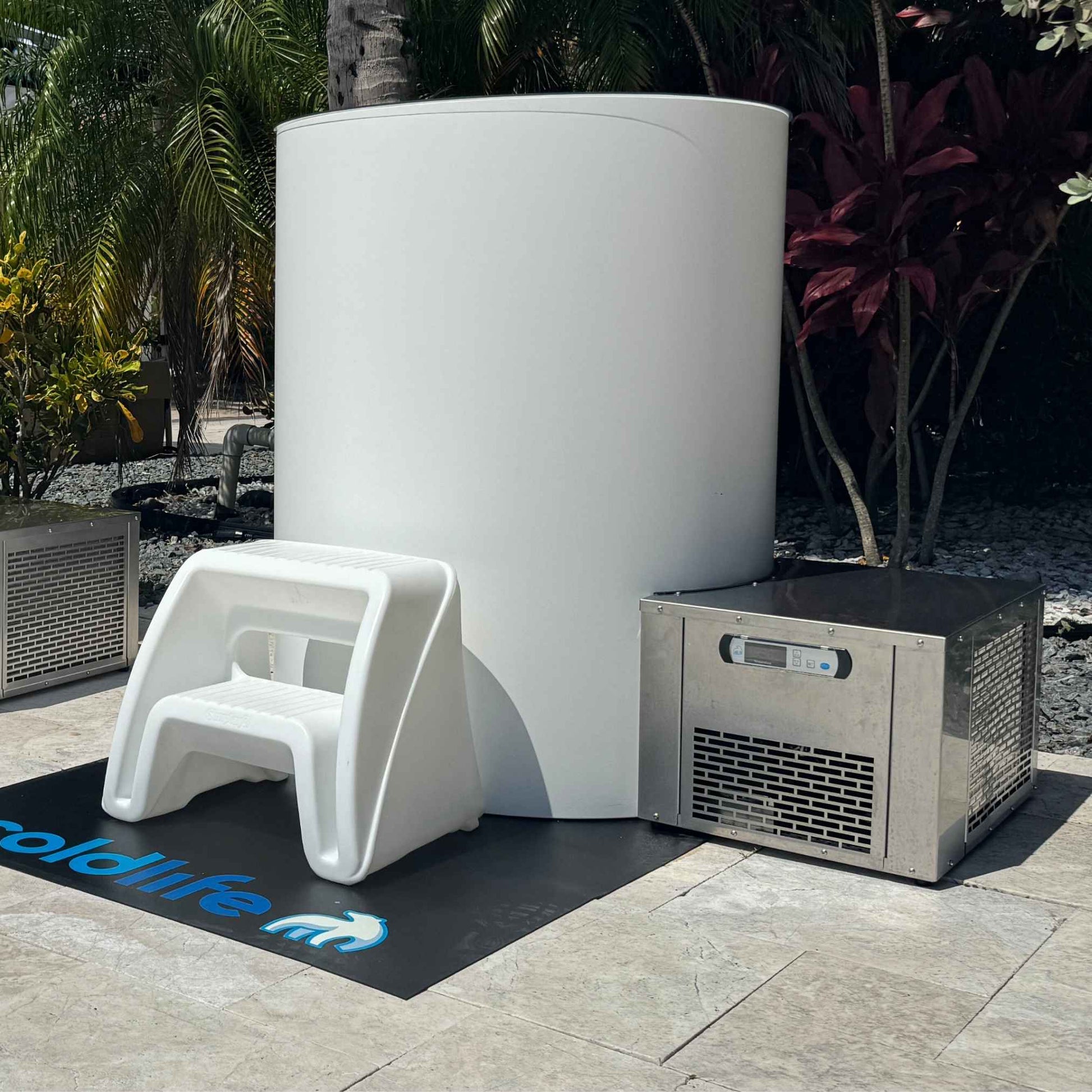 A white cylindrical unit next to a metallic cooling unit, with a small white step stool in front, all set on a black mat with the text coldlife visible, surrounded by tropical plants.