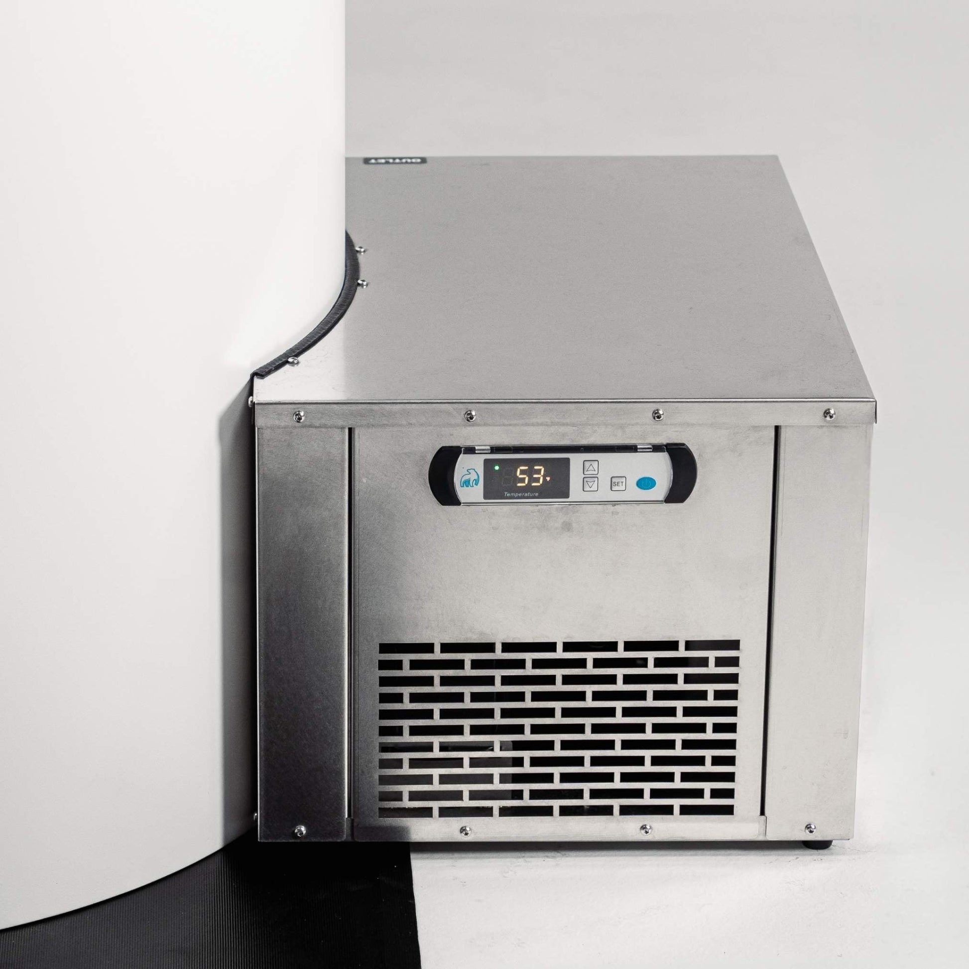 A stainless steel ice maker machine positioned in a corner, displaying a digital temperature reading of 53.3. The machine has a sleek design with a vented front panel and is set against a white wall and black floor.