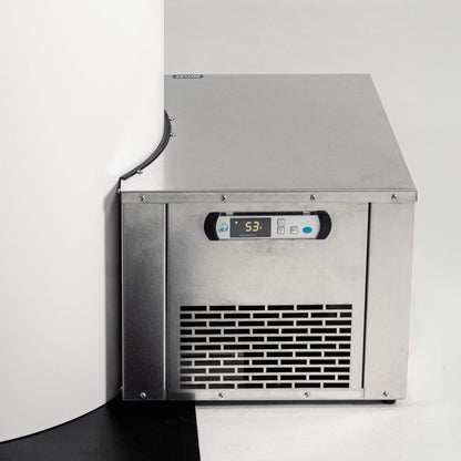 A stainless steel ice maker machine positioned in a corner, displaying a digital temperature reading of 53.3. The machine has a sleek design with a vented front panel and is set against a white wall and black floor.