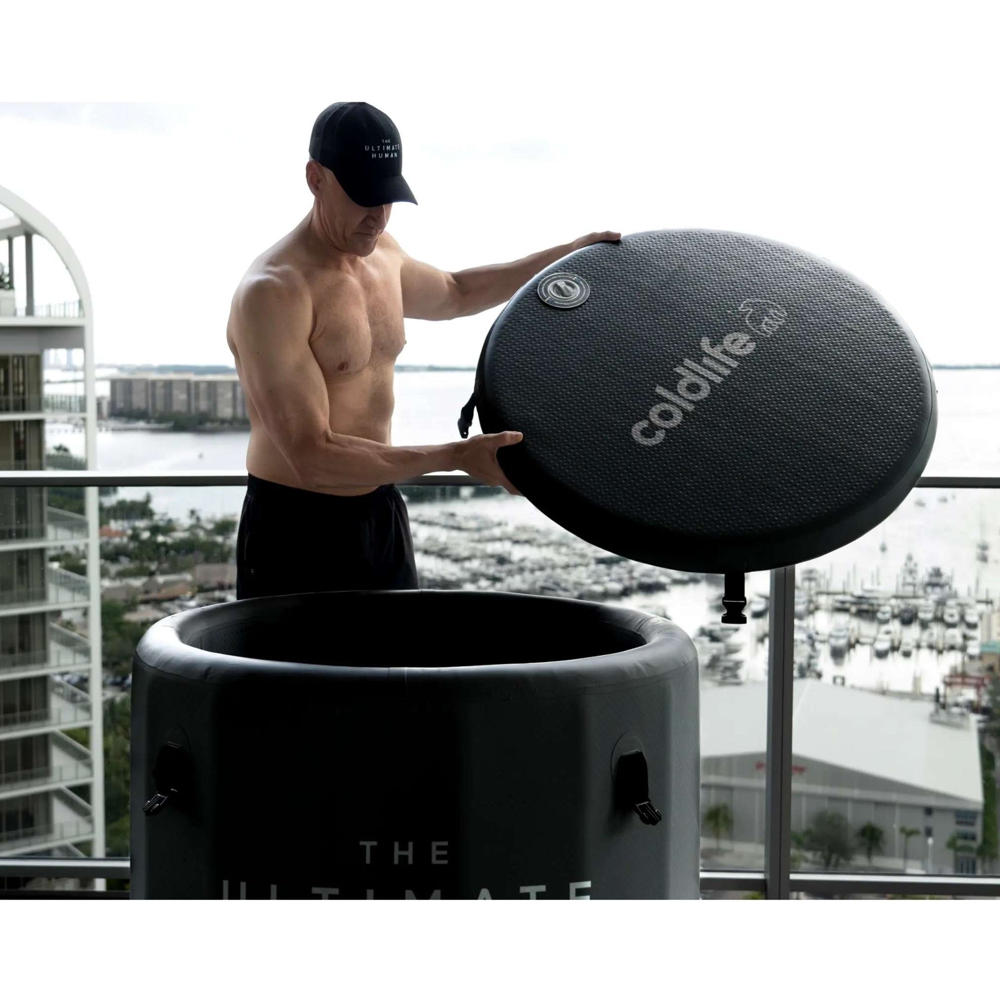 A shirtless man in athletic shorts is lifting the lid off a cold plunge tub labeled coldlife on a balcony, with a view of a marina and cityscape in the background.