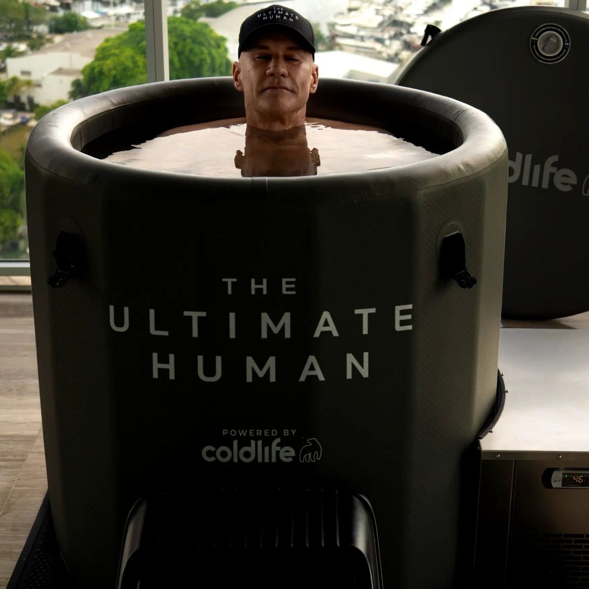 A person sitting in a cold therapy tub labeled 'THE ULTIMATE HUMAN' powered by coldlife, with a serene expression and a hat, surrounded by a modern interior.