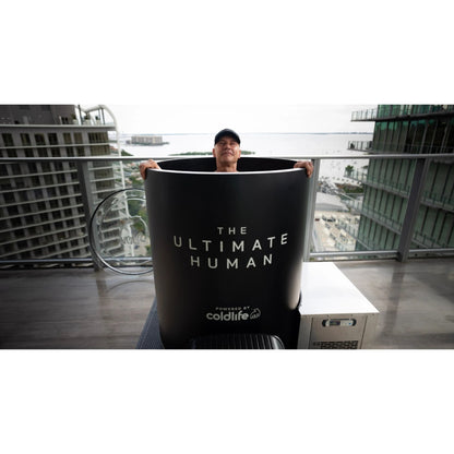 A person sitting in a large black tub labeled THE ULTIMATE HUMAN and powered by coldlife, with a cityscape view in the background.