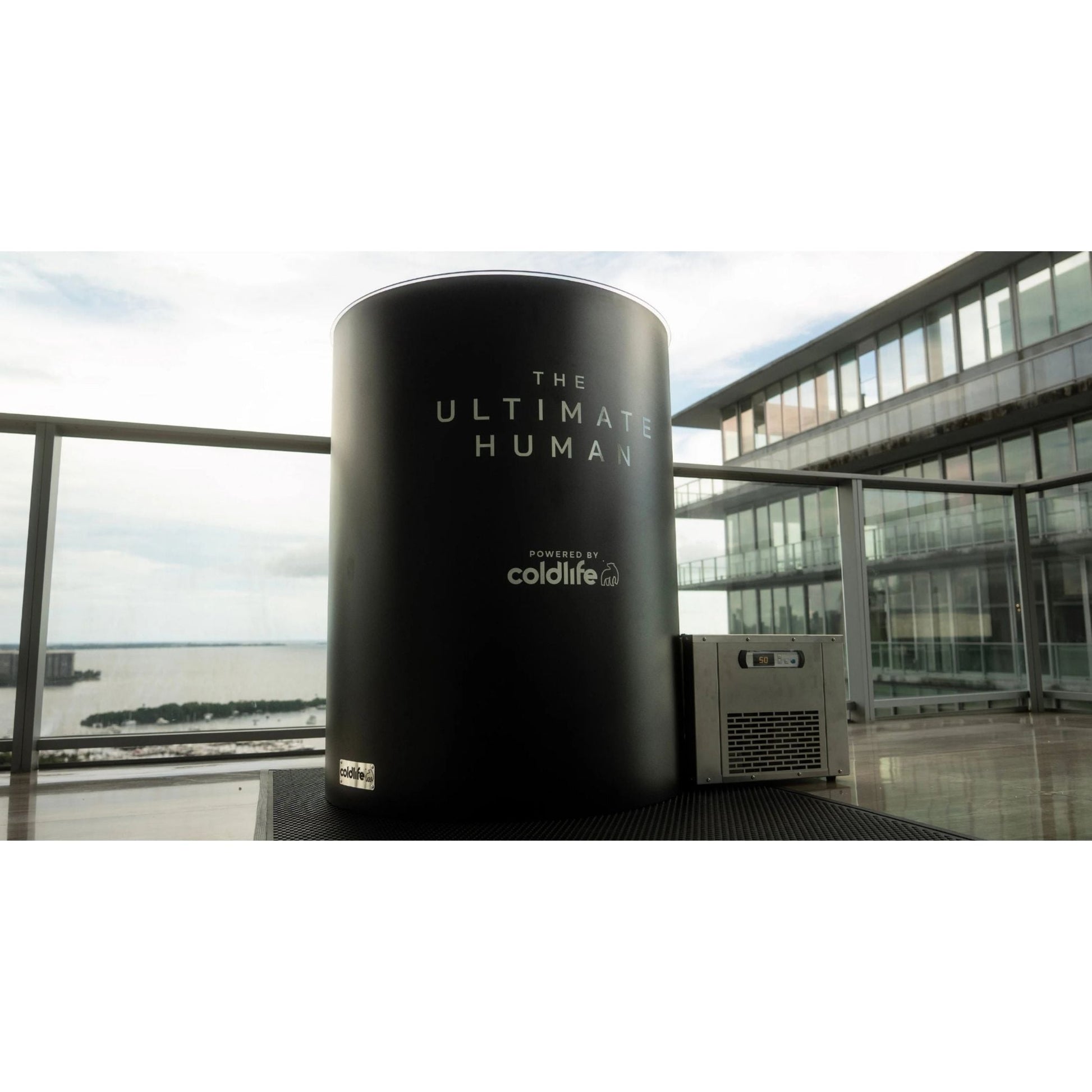 A black cylindrical display with the text THE ULTIMATE HUMAN and powered by coldlife on it, situated on a balcony with a view of the water and modern buildings in the background.
