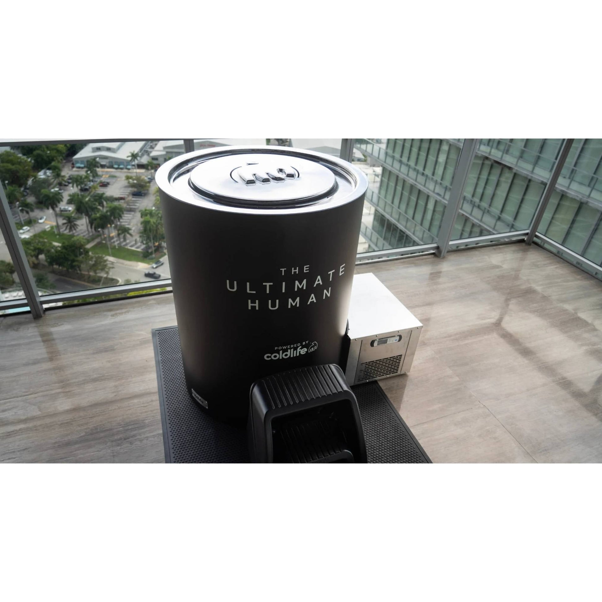 A modern, cylindrical device labeled THE ULTIMATE HUMAN powered by coldlife, placed on a sleek surface in a well-lit indoor space with a view of greenery outside.