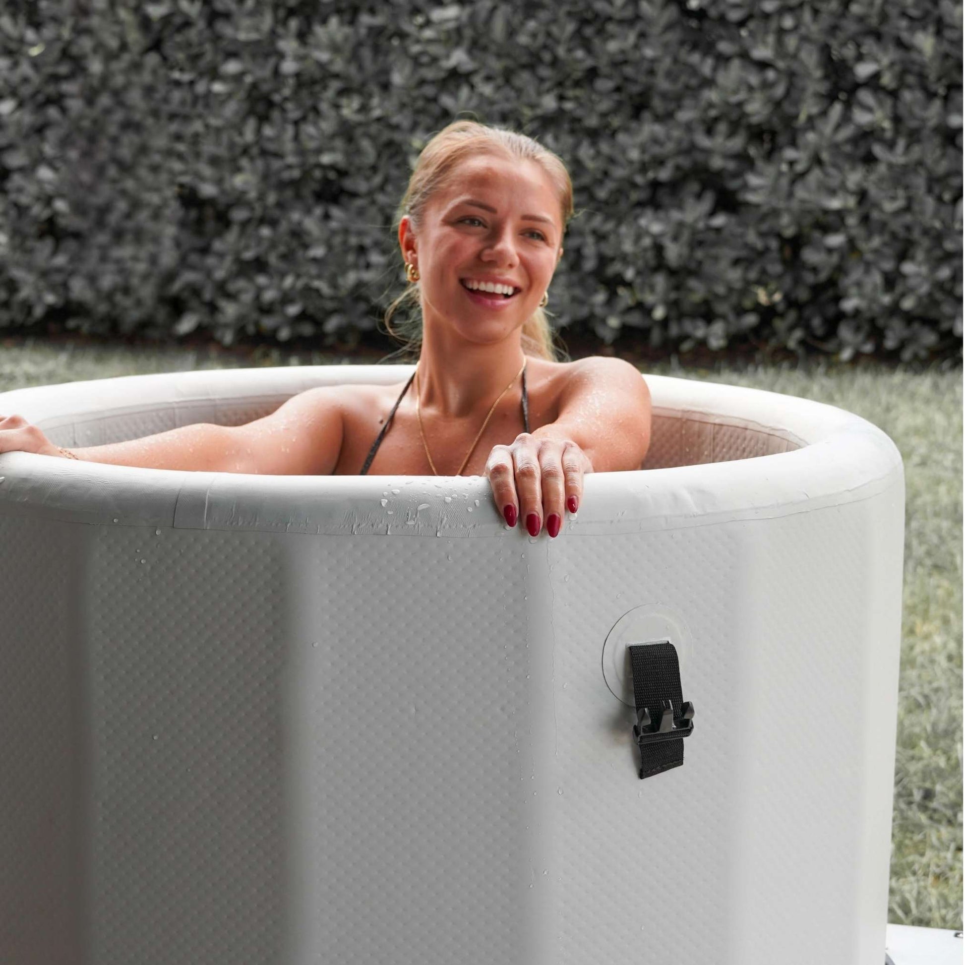 A woman with blonde hair smiles while relaxing in a white inflatable hot tub, surrounded by greenery. Water droplets are visible on the tub's surface.