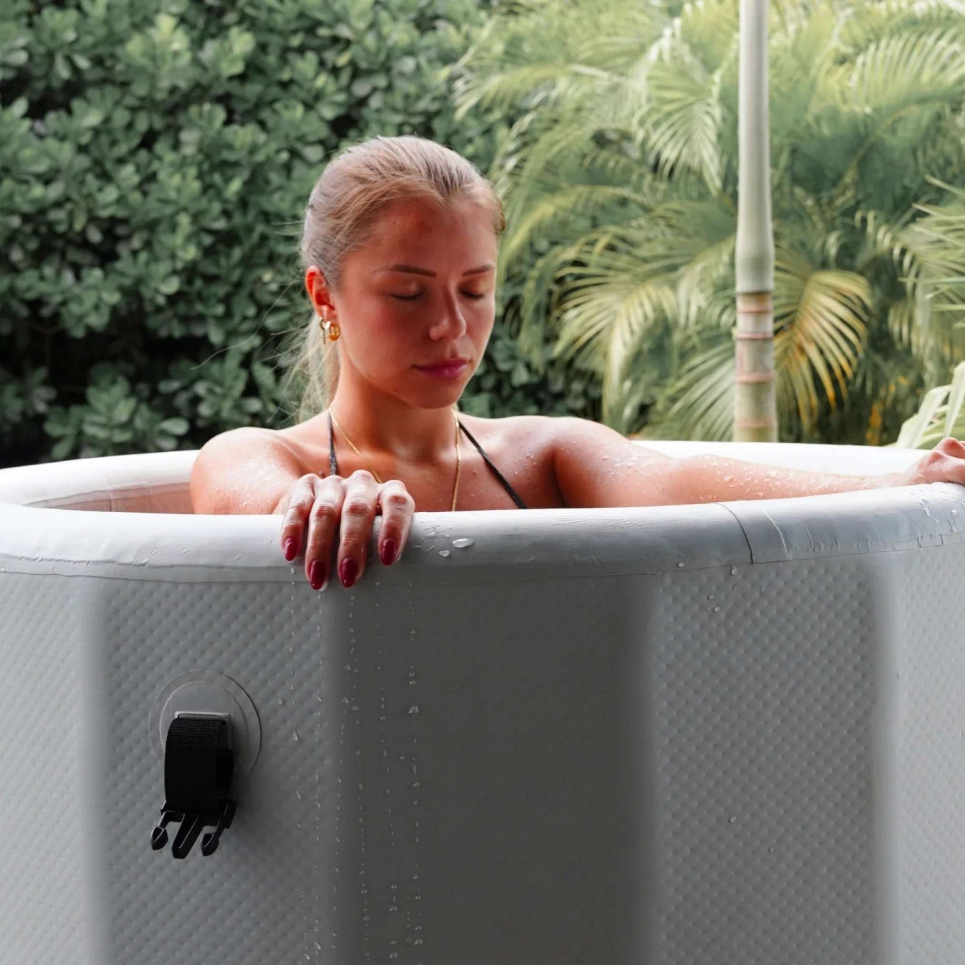 A woman with her eyes closed relaxes in a hot tub, surrounded by lush greenery. Water droplets glisten on her skin as she leans back, enjoying the serene atmosphere.