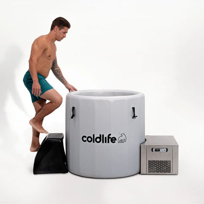 A man in swim trunks is stepping onto a black step stool to enter a gray cold plunge tub labeled coldlife, with a cooling unit beside it.
