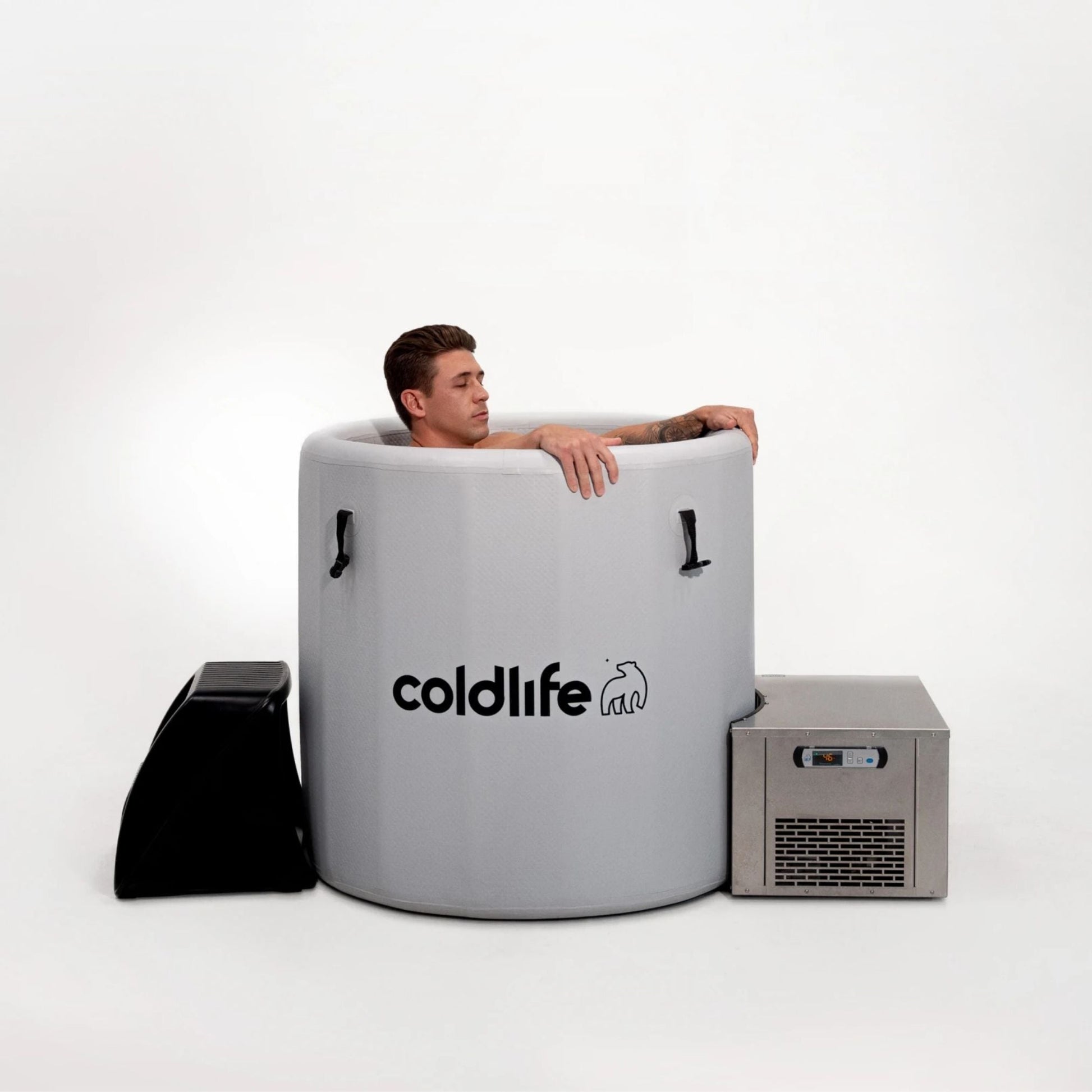 A man relaxes in a circular cold plunge tub branded with coldlife, next to a cooling unit, with a minimalist background.