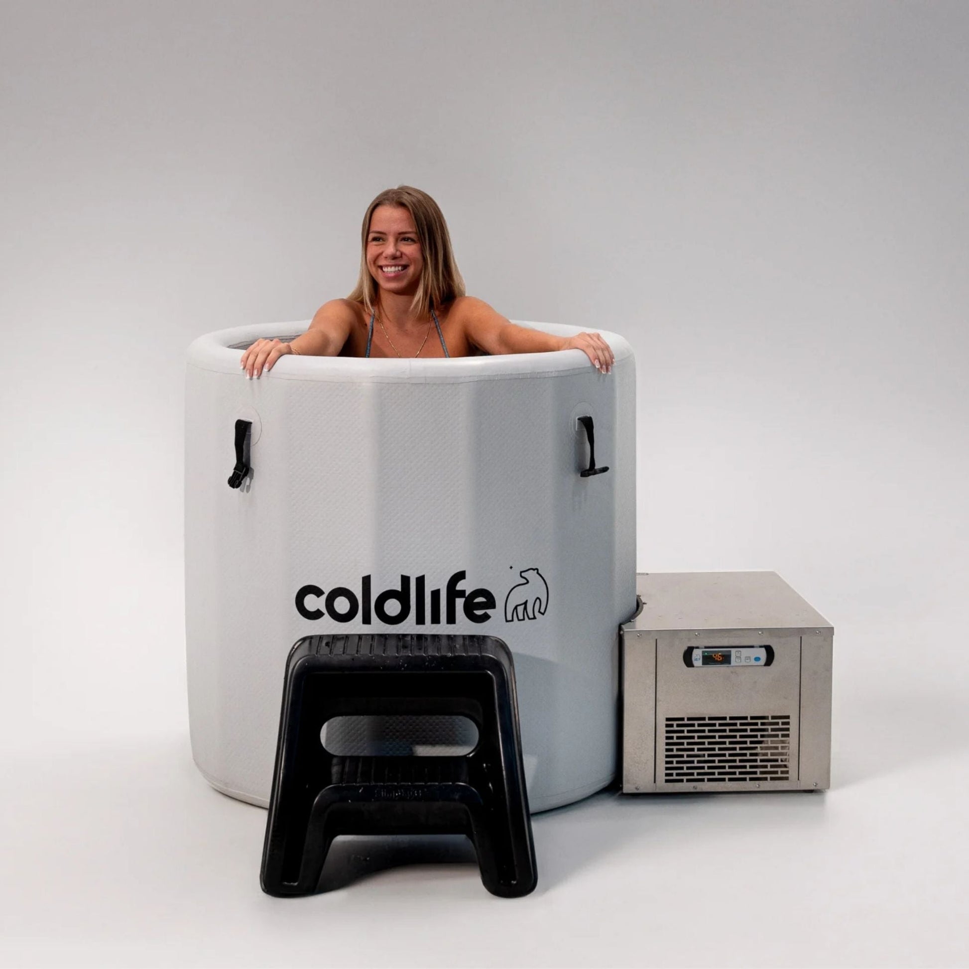 A smiling woman sitting in a cold therapy tub branded with 'coldlife,' next to a cooling unit and a small black stool, against a plain background.