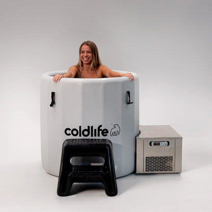 A smiling woman sitting in a cold therapy tub branded with 'coldlife,' next to a cooling unit and a small black stool, against a plain background.