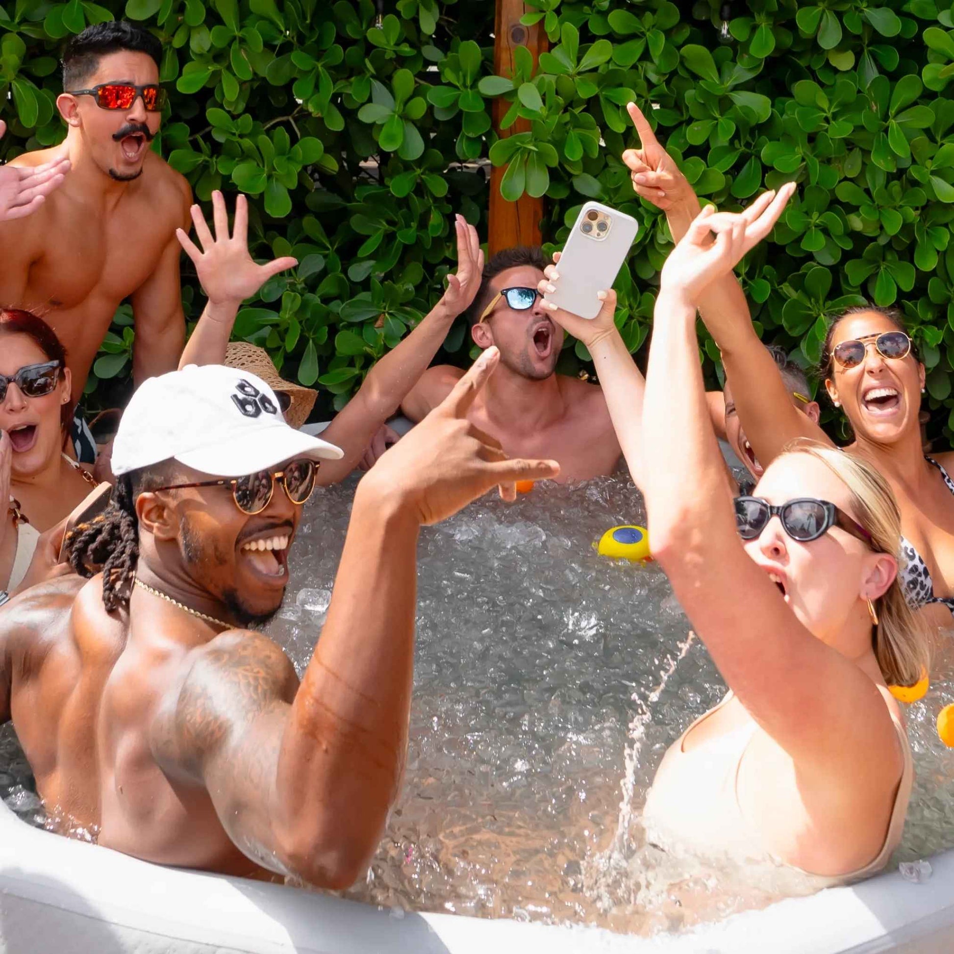 A lively group of people enjoying a fun moment in a hot tub, with hands raised and smiles all around, surrounded by lush greenery.