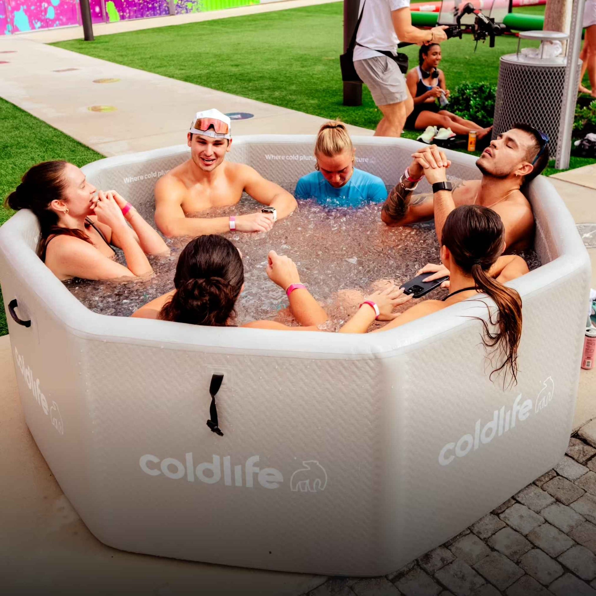 A group of six people enjoying a conversation while sitting in an inflatable hot tub branded coldlife, surrounded by a green lawn and colorful background decorations.