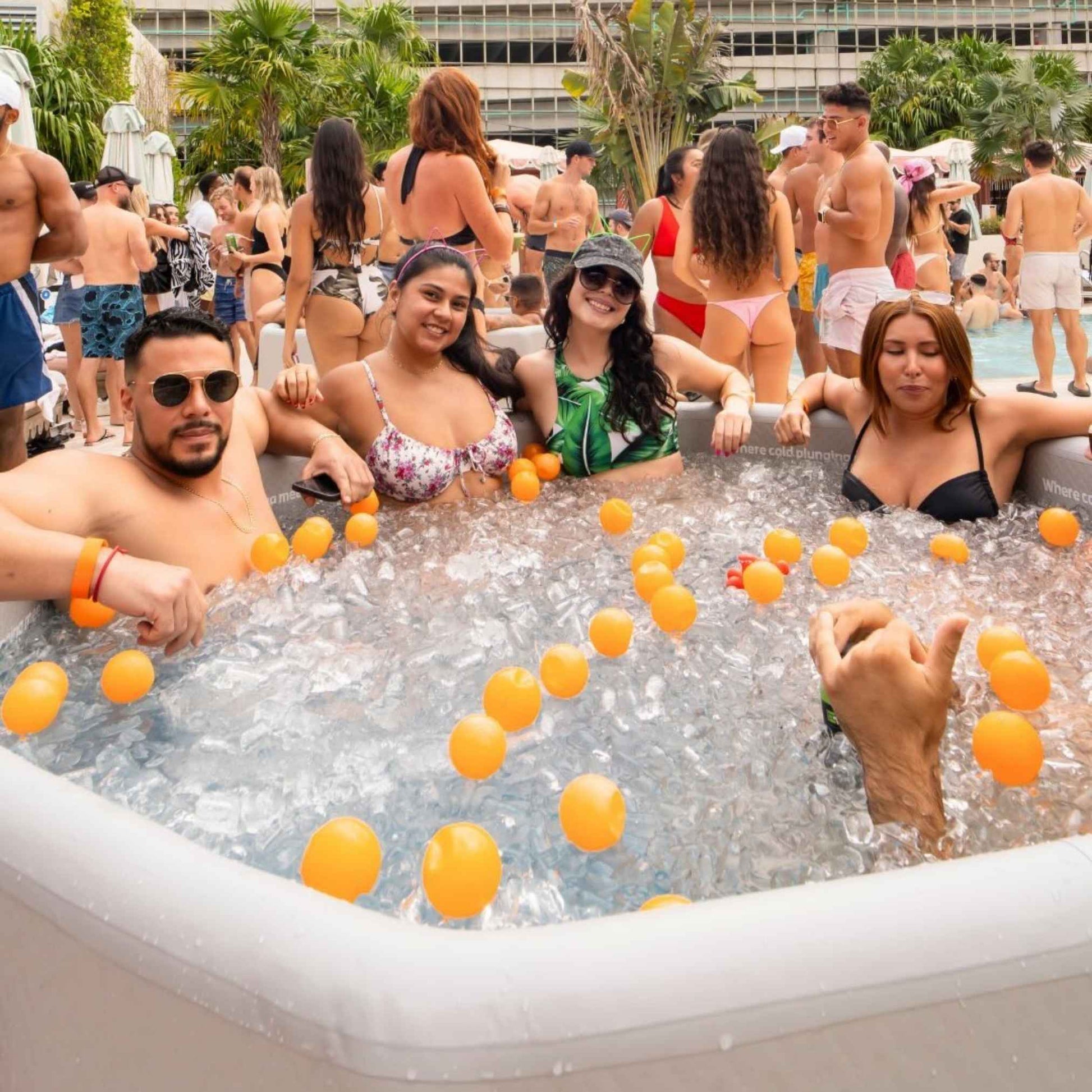 A group of four people enjoying a hot tub filled with ice and orange ping pong balls at a lively pool party, with many others socializing in the background.