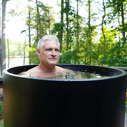 A man with short gray hair relaxing in a black tub filled with water, surrounded by lush green trees and a serene outdoor setting.