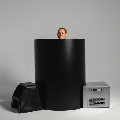 A woman’s head is visible above a large black cylindrical structure, with a black stool on the left and a silver freezer unit on the right, set against a light gray background.