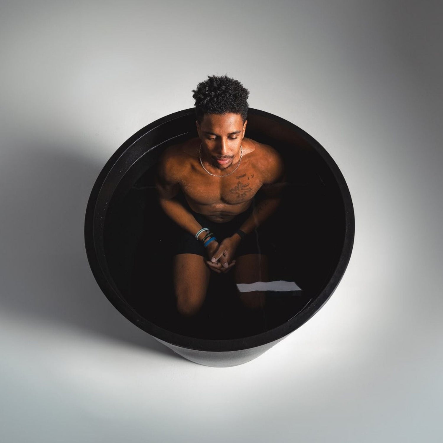 A person sitting in a black tub filled with water, with a calm expression, wearing black shorts and bracelets, surrounded by a minimalistic background.