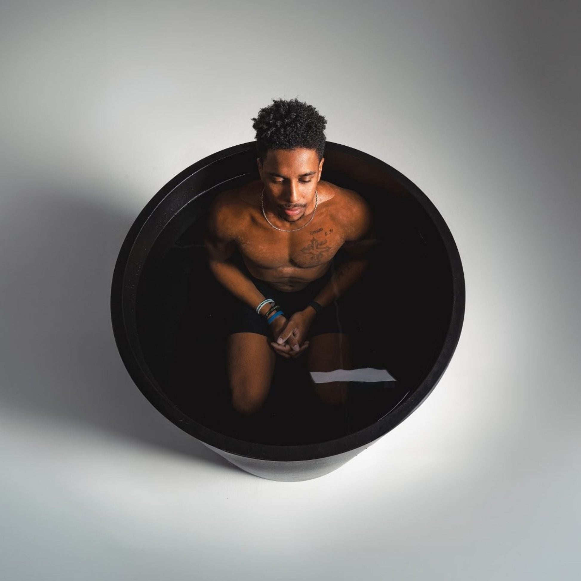 A person sitting in a black tub filled with water, with a calm expression, wearing black shorts and bracelets, surrounded by a minimalistic background.